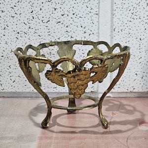 May include: A gold metal decorative stand with a round top and three legs. The stand features a grapevine design with grapes.