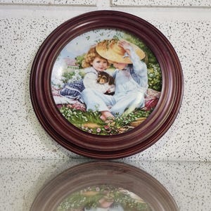 May include: A round decorative plate with a brown wooden frame. The plate features a pastoral scene of two children and a puppy. The children are wearing light blue clothing and sitting in a field of flowers. The plate is approximately 20 cm in diameter.