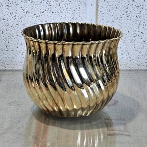 May include: A gold-coloured decorative vase with a wavy, vertical ribbed design. The vase has a wide, open top with a scalloped edge. The surface reflects light, creating highlights and shadows.
