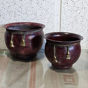 May include: Two dark red metal planters with gold trim and tassels. The planters are on a patterned surface.