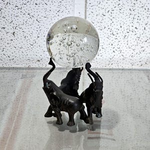 May include: A clear glass sphere with air bubbles is held aloft by a bronze-coloured elephant sculpture. The sculpture depicts three elephants with raised trunks. The elephants appear to be supporting the glass sphere, creating a unique display.