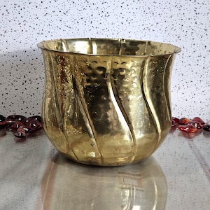 Peut inclure: Un pot en laiton martelé avec un design cannelé. Le récipient doré a une large ouverture et une surface texturée. Le pot est posé sur une surface réfléchissante.