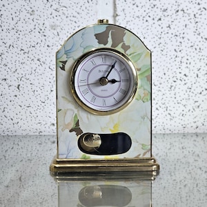 Peut inclure: Une horloge à quartz Bulova avec un motif floral. Le cadran blanc affiche des chiffres romains et des détails dorés. Le boîtier est orné d'un motif floral blanc, jaune et vert. Elle possède un pendule et un socle doré. Dimensions: environ 15 cm de hauteur.