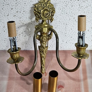 May include: A vintage brass wall sconce with two arms, each holding a light fixture. The sconce features floral and leaf detailing. Two gold-colored cylindrical sleeves are included. The sconce is designed for two light bulbs.