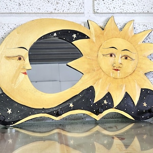 Puede incluir: Espejo de pared de madera con diseño de sol y luna. El sol es amarillo con una cara sonriente, y la luna tiene forma de media luna con una cara. El diseño incluye detalles en negro con estrellas blancas y pequeños ganchos.