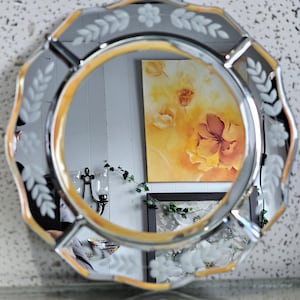 Może przedstawiać: Ośmiokątne lustro ze złotą obwódką i grawerowanymi wzorami kwiatów i liści. Lustro odbija obraz z żółtymi i pomarańczowymi kwiatami. Dekoracyjny element wystroju wnętrz.