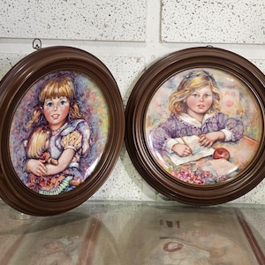 May include: Two vintage decorative plates in brown oval frames. Each plate features a painted portrait of a young girl. One girl holds a doll, and the other is writing. The plates have a glossy finish and are mounted on a wall.