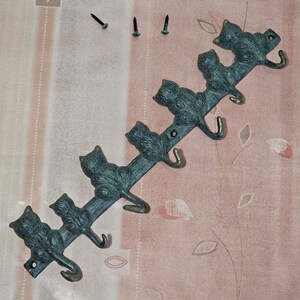 Könnte beinhalten: Ein Schlüsselhalter aus Metall mit sechs katzenförmigen Haken. Die Katzen sind grün und bronzefarben und schauen nach links. Der Halter ist auf einer rosa-weißen gemusterten Oberfläche montiert.