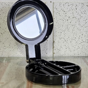 Peut inclure: Un miroir de courtoisie noir de style Art déco avec un miroir rond pivotant et une base à compartiments. Le miroir a un bord perlé décoratif. La base a une forme circulaire avec trois compartiments.