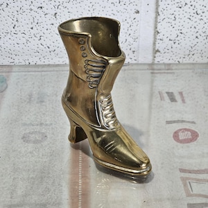 May include: A gold-colored brass boot-shaped vase with a high heel. The boot has a decorative design on the top and side.