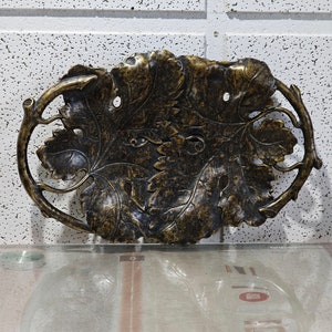 May include: A decorative metal tray with a grapevine design. The tray is made of a dark metal with a gold finish. The tray is oval shaped and has a raised rim.