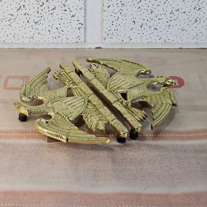 May include: A gold-colored metal hot pad shaped like two eagles with their wings spread. The eagles are facing each other and have a decorative design on their chests.