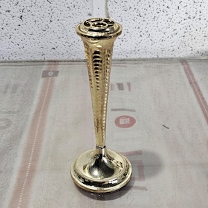 Peut inclure: Un soliflore doré orné, avec une base ronde et une tige élancée et cannelée. Le sommet présente un motif décoratif ouvert. Le vase est en métal et a une finition brillante. Le vase mesure environ 20 cm de haut.