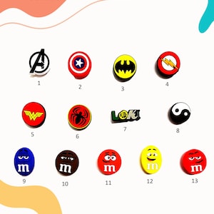 May include: Thirteen colourful shoe charms featuring various superhero and cartoon characters. The charms are arranged in three rows of four, with one charm in the bottom right corner. The charms include characters such as Spiderman, Wonder Woman, Batman, Captain America, the Avengers logo, the Flash, Loki, Yin Yang, and M&M's.