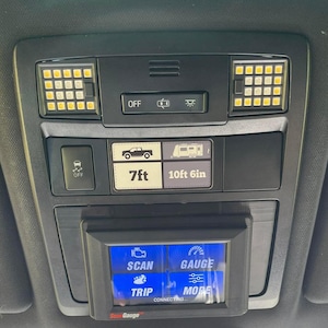 May include: A black car interior console with a digital display and various buttons. The display shows "SCAN", "GAUGE", "TRIP", and "MORE". Above the display are buttons labelled "OFF", and icons for lights and a vehicle. The console also has LED lights.