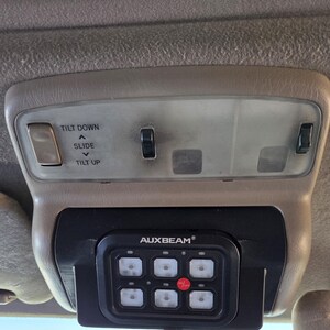 May include: Tan interior car console with overhead lights and an AUXBEAM control panel. The console has buttons labeled "TILT DOWN", "SLIDE", and "TILT UP". The AUXBEAM panel has six square buttons and a red button labeled "on/off".