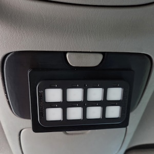 May include: A black Switch-Pros panel with eight illuminated square buttons, mounted on a vehicle's gray interior. The panel is rectangular and has the text "SWITCH-PROS" printed on the side. The buttons are arranged in two rows of four.