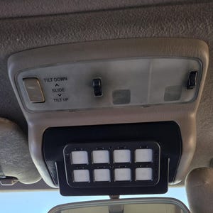 May include: Tan interior car console with a black control panel featuring eight illuminated square buttons. Above, a light fixture with switches labeled "TILT DOWN", "SLIDE", and "TILT UP". A rearview mirror is visible below.