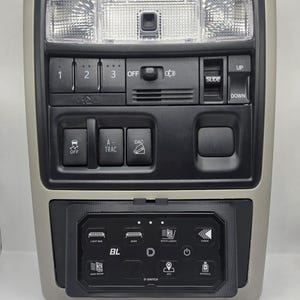 May include: A beige and black overhead console with multiple buttons and switches. Features include light controls, a sunroof slider, and various function buttons. A lower panel displays additional controls with the letters "BL" and "D-SWITCH".