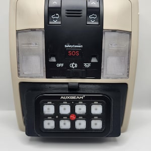 May include: A beige overhead console with a black control panel featuring buttons for sunroof, SOS, and lights. A black AUXBEAM device with nine buttons is attached to the bottom of the console.