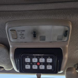 May include: Tan interior car console with overhead lights and a black AUXBEAM control panel. The panel has eight illuminated square buttons and the text "AUXBEAM". Above are controls labeled "TILT DOWN", "SLIDE", and "TILT UP".