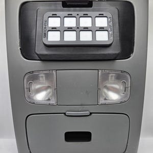 May include: A gray automotive overhead console with a black switch panel. The switch panel has eight illuminated white switches and the text "SWITCH-PROS" and "RCR-TOUCH-09". Below are two clear dome lights and a storage compartment.