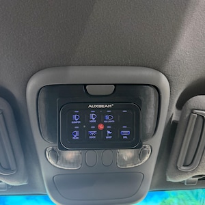 May include: A black Auxbeam switch panel mounted on a vehicle ceiling console. The rectangular device features six illuminated blue buttons with icons and labels for Bumper, Hood, Fog Lights, Rear, Rock, Whip, and DRL, plus a central red power button.