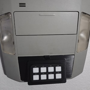 May include: A gray overhead console with a black switch panel attached. The switch panel has eight white square buttons. The console has two lights and a storage compartment.