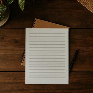 Puede incluir: Una hoja de papel blanco con líneas descansa sobre un cuaderno en espiral marrón sobre una superficie de madera oscura. Un bolígrafo negro está colocado cerca del papel. Una planta verde y una estera tejida son visibles en el fondo, sugiriendo un espacio de trabajo.