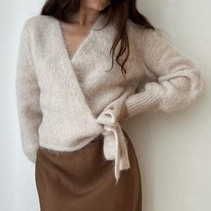 May include: A light beige wrap sweater with long sleeves and a tie closure. The sweater has a soft, fuzzy texture and a V-neckline. The bottom half of the image shows a brown skirt.