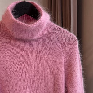 May include: A soft, pink turtleneck sweater with long sleeves hangs on a black hanger. The sweater is made of a fuzzy, knitted material. The turtleneck is folded over. The sweater is a solid color.