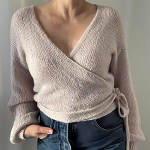 May include: A light pink wrap sweater with long sleeves and a V-neck. The sweater is tied at the waist with a matching fabric tie. The sleeves are cuffed at the wrists. The sweater is paired with blue jeans.