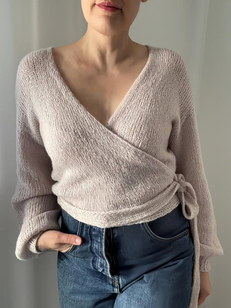 May include: A light pink wrap sweater with long sleeves and a V-neck. The sweater is tied at the waist with a matching fabric tie. The sleeves are cuffed at the wrists. The sweater is paired with blue jeans.