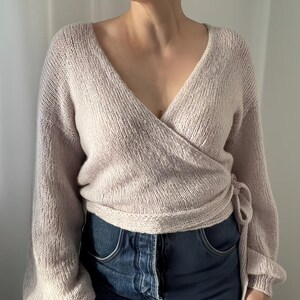 May include: A light beige wrap sweater with long sleeves and a V-neck. The sweater is tied at the waist and is paired with blue denim jeans. The knitwear has a soft, textured appearance, suggesting a cozy and comfortable feel.
