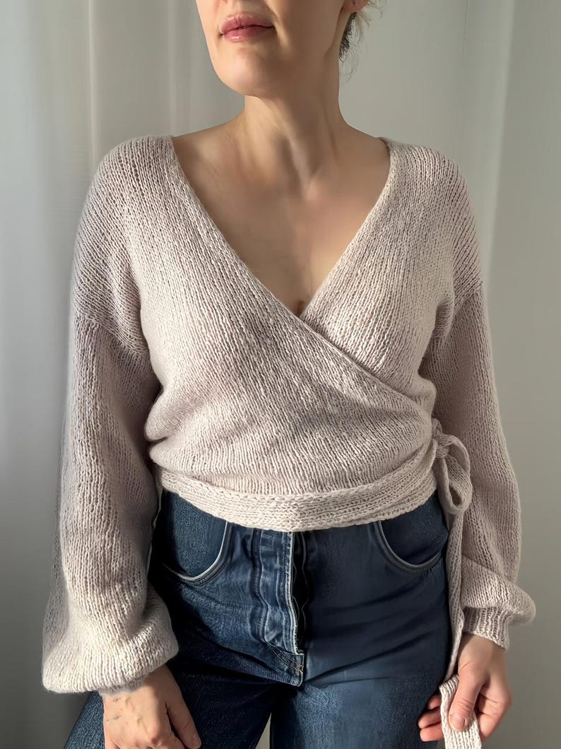May include: A light beige wrap sweater with long sleeves and a V-neck. The sweater is tied at the waist and is paired with blue denim jeans. The knitwear has a soft, textured appearance, suggesting a cozy and comfortable feel.