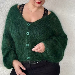 Puede incluir: Un c&aacute;rdigan verde oscuro y peludo con cuello en V y cierre de botones. El c&aacute;rdigan se lleva sobre una camiseta negra y unos vaqueros negros.