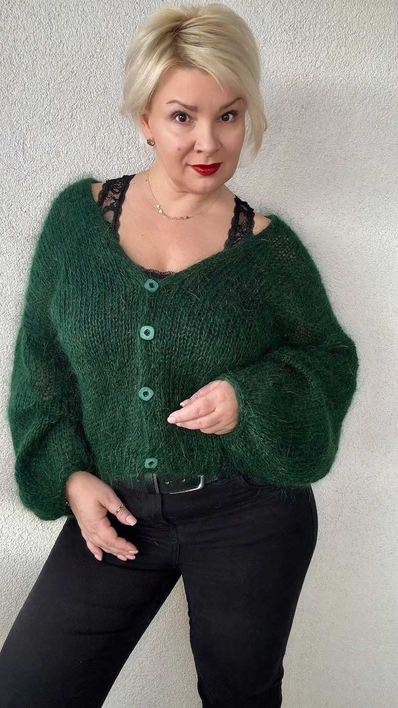 Puede incluir: Un c&aacute;rdigan verde oscuro y peludo con cuello en V y cierre de botones. El c&aacute;rdigan se lleva sobre una camiseta negra y unos vaqueros negros.