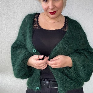 Puede incluir: Una chaqueta de punto verde oscuro y peluda con dos botones. La chaqueta se lleva sobre un top negro de tirantes y unos vaqueros negros.