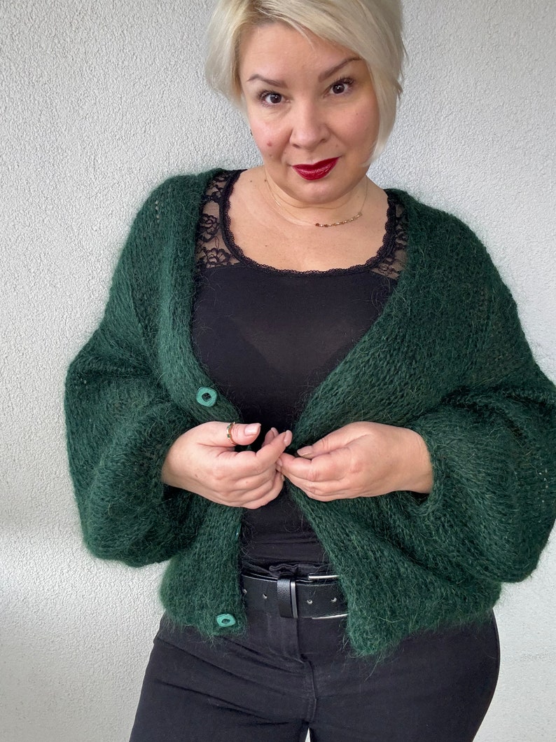 Puede incluir: Una chaqueta de punto verde oscuro y peluda con dos botones. La chaqueta se lleva sobre un top negro de tirantes y unos vaqueros negros.