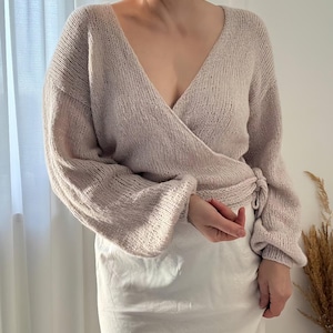 May include: A light beige wrap sweater with a deep V-neck and long sleeves. The sweater is tied at the waist and has a soft, knitted texture. The sleeves are slightly puffed at the shoulders. The model is wearing a white skirt.