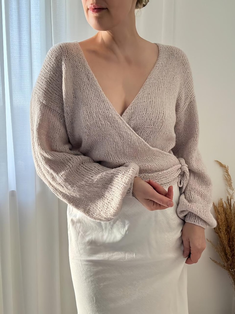 May include: A light beige wrap sweater with a deep V-neck and long sleeves. The sweater is tied at the waist and has a soft, knitted texture. The sleeves are slightly puffed at the shoulders. The model is wearing a white skirt.