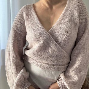 May include: A light pink wrap sweater with a deep V-neck and long sleeves. The sweater is made of a soft, knitted material and has a ribbed waistband and cuffs. The style is a cozy, feminine design.