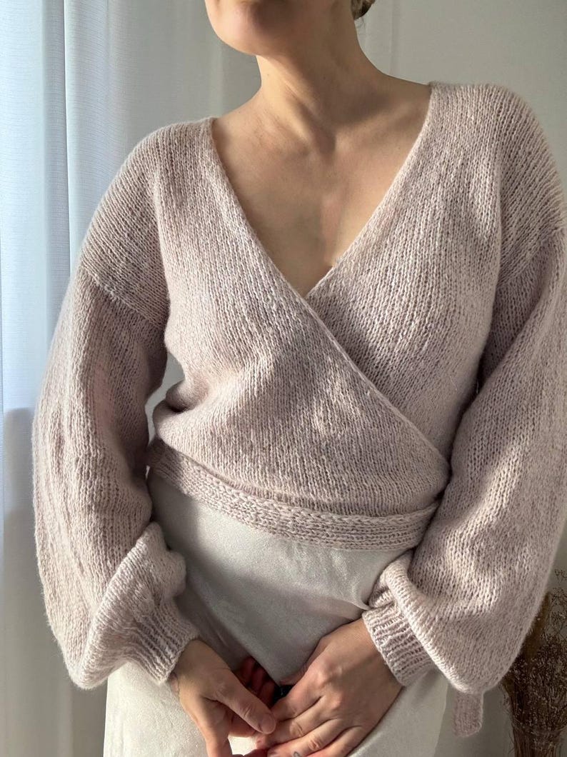 May include: A light pink wrap sweater with a deep V-neck and long sleeves. The sweater is made of a soft, knitted material and has a ribbed waistband and cuffs. The style is a cozy, feminine design.