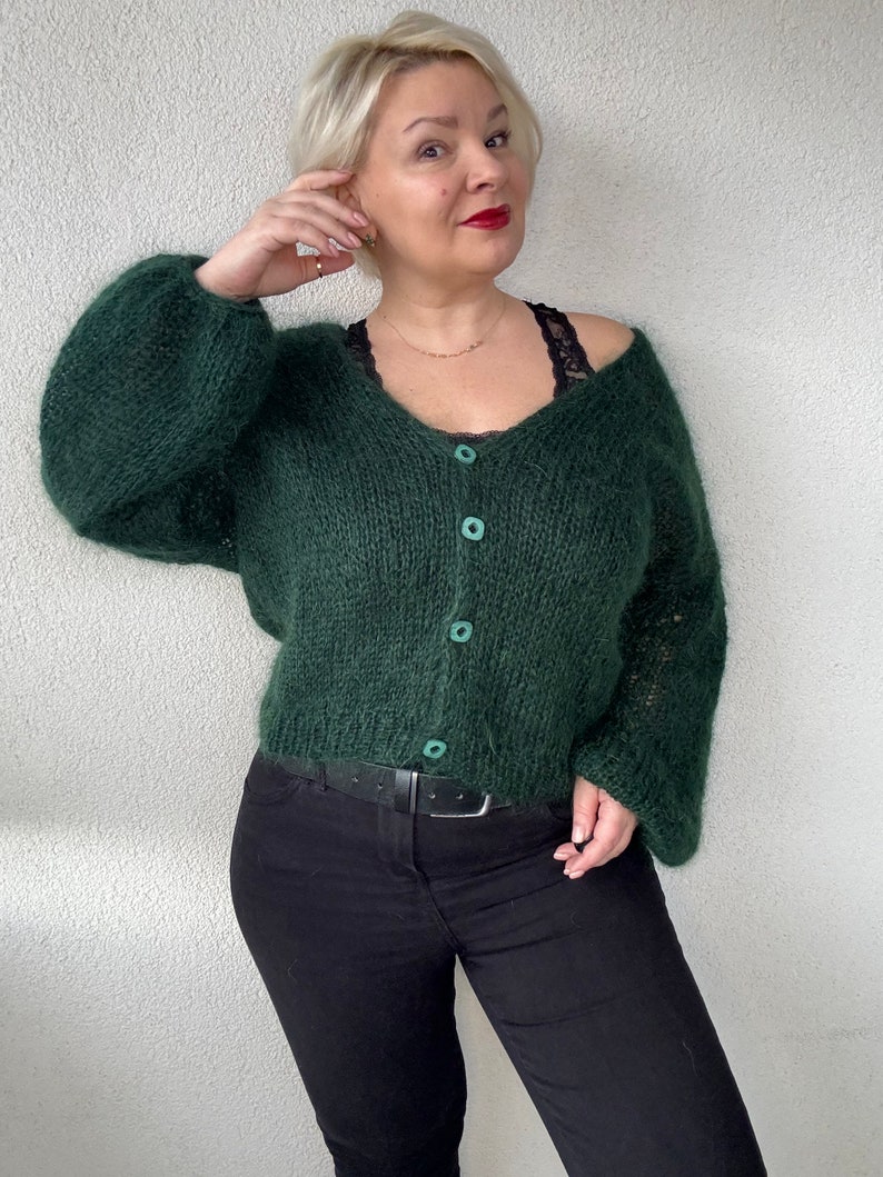 Puede incluir: Un c&aacute;rdigan de color verde oscuro y peludo con cuello en V y cierre de botones. El c&aacute;rdigan tiene mangas largas y un corte holgado.