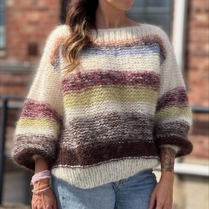 Può includere: Un maglione oversize a righe orizzontali color crema, marrone, giallo, viola e blu. Il maglione ha una scollatura ampia e maniche voluminose. La maglia appare morbida e strutturata, perfetta per la stagione fredda.