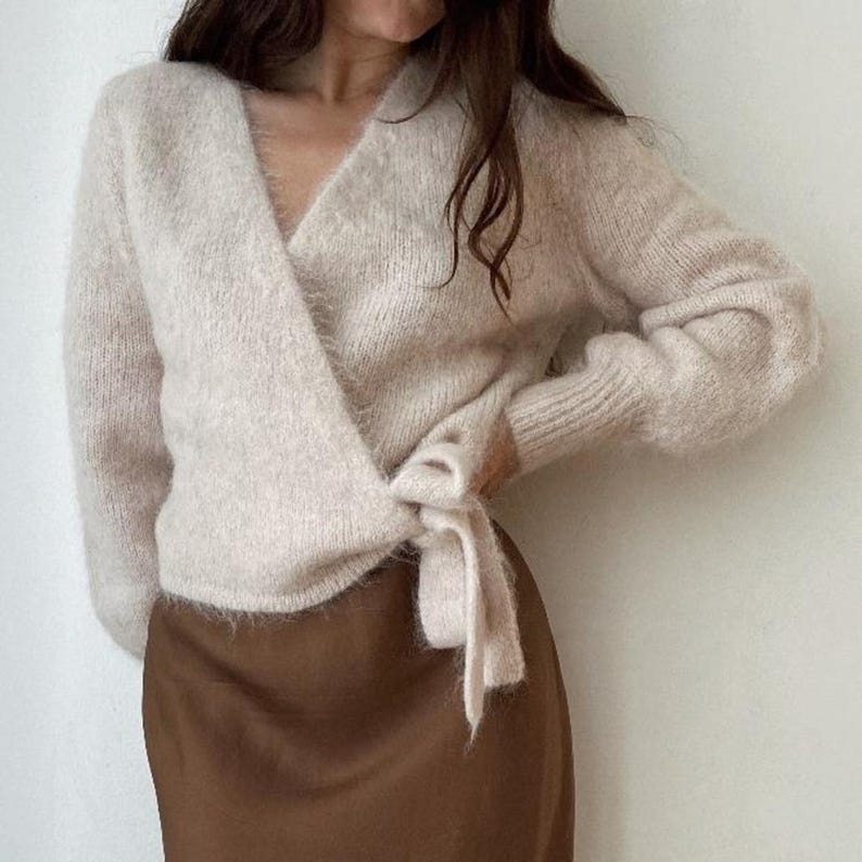May include: A light beige wrap sweater with long sleeves and a tie closure. The sweater has a soft, fuzzy texture and a V-neckline. The bottom half of the image shows a brown skirt.