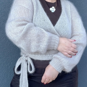 May include: A light gray, wrap-style sweater with long sleeves and a tie closure at the waist. The sweater is made of a soft, fuzzy material. The person in the image is wearing a dark brown top and black pants.