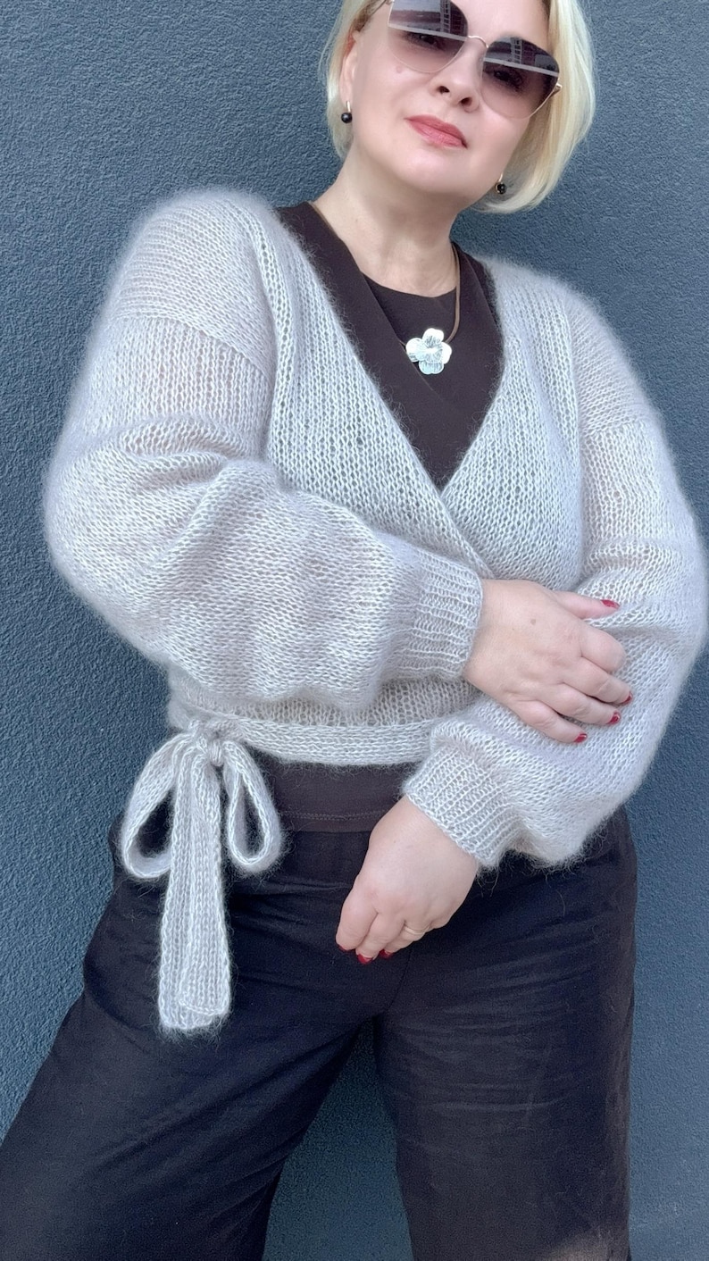 May include: A light gray, wrap-style sweater with long sleeves and a tie closure at the waist. The sweater is made of a soft, fuzzy material. The person in the image is wearing a dark brown top and black pants.