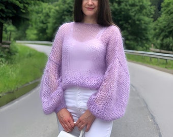 Puede incluir: Un suéter de punto de color lila claro con un corte holgado y mangas largas abullonadas. El suéter está hecho de un hilo delicado y aireado y tiene una apariencia ligeramente transparente.
