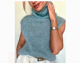 Puede incluir: Un suéter sin mangas de cuello alto en color azul claro con una textura de punto grueso y abierto. La prenda está confeccionada con un hilo suave y esponjoso, combinada con pantalones blancos que tienen una hebilla plateada visible.
